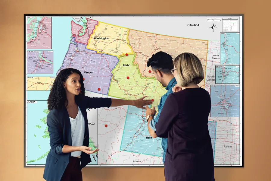 Three colleagues discussing a Northwest US wall map pinned with red markers across Washington, Oregon, Idaho and Utah, with city inset maps along both edges.